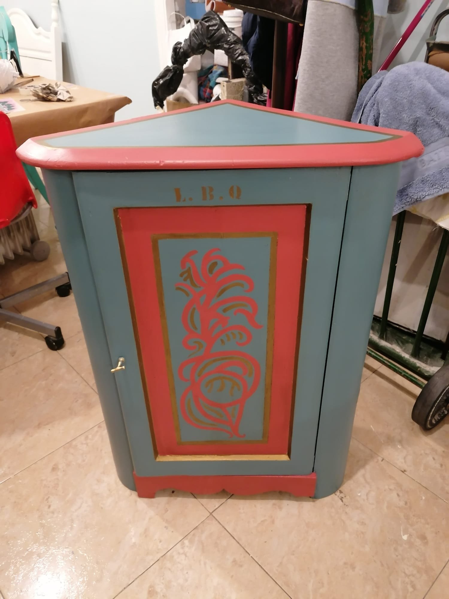 Armario esquinero de madera pintado en azul turquesa y rojo coral, con un diseño decorativo en la puerta y las letras "L.B.Q." grabadas en la parte superior.